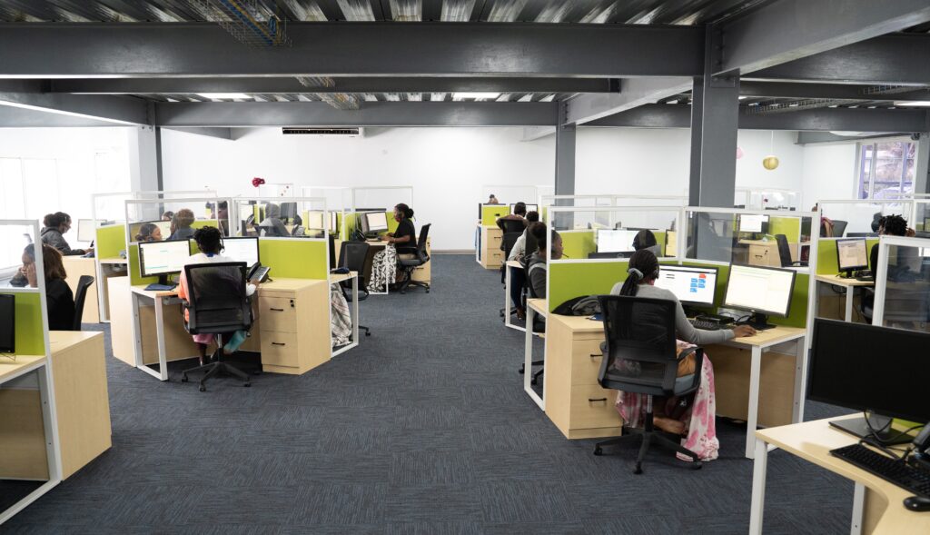 Image featuring itel's Saint Lucia site. The COVID-ready production floor features wide walkway areas between y-shaped desk configurations, with high plexiglass partitions that allow coworkers to communicate safely. 