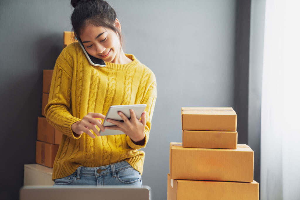 Image of young, happy Asian woman on her cell phone and tablet, surrounded with packages