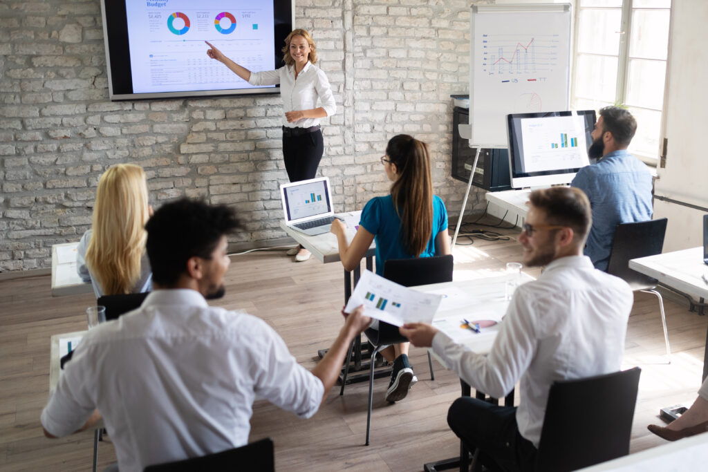 Image of an in-office training class. A teacher is presenting a slideshow to several trainees, sitting in seats, in front of computers